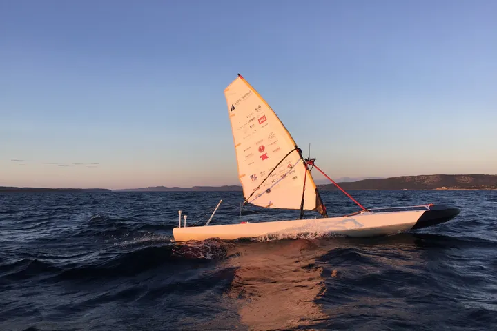 Ada, UBC Sailbot’s autonomous sailboat, sailing at sunset under a sail covered in sponsor logos.